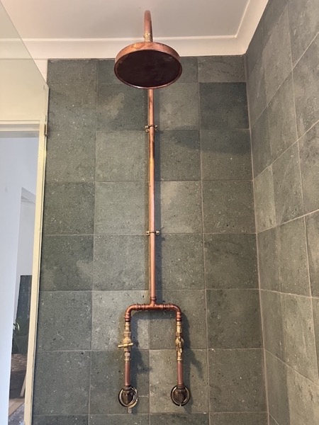 An exposed copper shower with a copper rose and brass handles on slate tiles