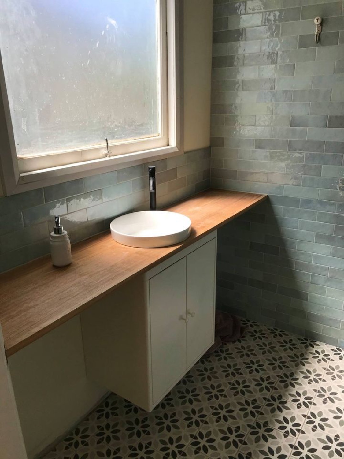A timber vanity top with a round white basin and a black tall mixer against sage subway tiles
